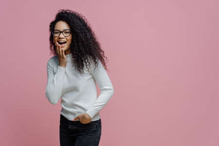 Photo of slim cheerful African American woman with curly hair, smiles happily, being in good mood, wears white t shirt and jeans, keeps hand in pocket, has slim figure, isolated on beige backgroundの写真素材
