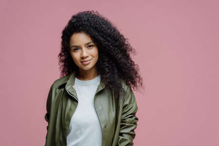 Horizontal shot of happy African American woman keeps hand in pocket, laughs as hears funny anecdote, spends free time in merry company, concentrated down, isolated on pink background, copy spaceの写真素材