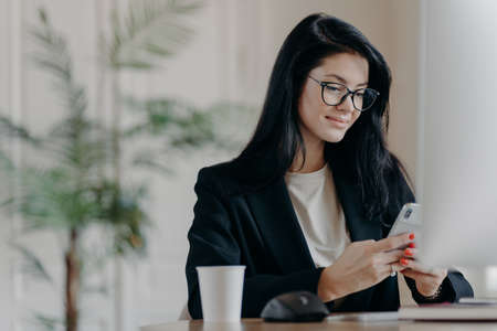 Satisfied business lady uses smartphone, poses at work place in front of computer screen, cup of tasty beverage, sends messages during break, poses at contemporary office. Technology and work conceptの写真素材