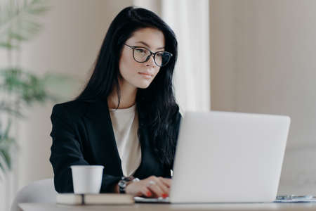 Concentrated brunette woman IT specialist in eyewear searches information using internet, keyboards on laptop computer, drinks coffee, has serious expression. Business and freelance concept.の写真素材