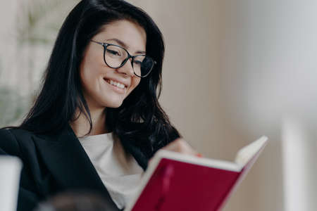 Cheerful dark haired woman creats do list with targets, holds notebook, writes organization plan, wears spectacles, dressed in formal outfit, smiles pleasantly. Autodidact and education conceptの写真素材