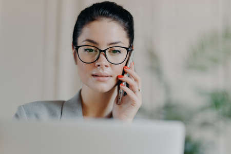 Horizontal shot of dark haired experienced woman with serious expression, makeup, talks via mobile phone, concentrated into screen, calls business partner to talk about working issues, office interiorの写真素材