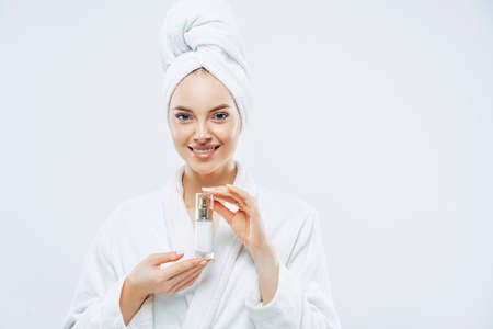 Beautiful spa young woman with healthy fresh skin applies anti aging lotion or cosmetic cream, uses day moisturizer, stands indoor, dressed in bath robe and towel, takes shower before going outの写真素材