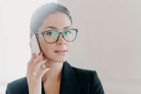 Close up shot of attractive brunette woman talks on mobile phone, looks seriously and self confident, makes call to banking service, has red manicure, wears optical glasses, formal black suitの写真素材