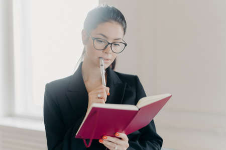Serious skilled Caucasian woman bank financial analyst examines business information, makes ideas how to invest money, writes down notes in notepad, wears spectacles and formal suit, poses indoorの写真素材
