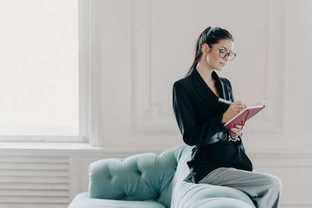 Attracative successful businesswoman in elegant clothes records information in notepad, sits on back...