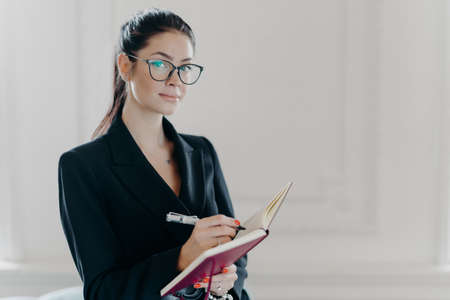 Serious elegant female teacher in black formal suit plans her working schedule, writes down information or some ideas in organizer, wears transparent glasses, poses indoor, white background.の写真素材
