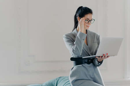Elegant professional woman banker concentrated at screen of laptop computer, dressed in formal wear,...