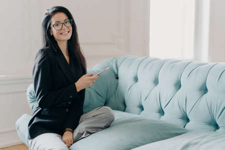 Happy business lady in official clothes, sits on comfortable sofa, uses mobile phone, reads news via network, searches information in cellular, going to phone partner, explores online projectの写真素材