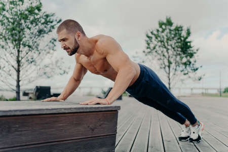 Confident muscled sportsman in sport wear doing push up outdoors, works on biceps and trains...