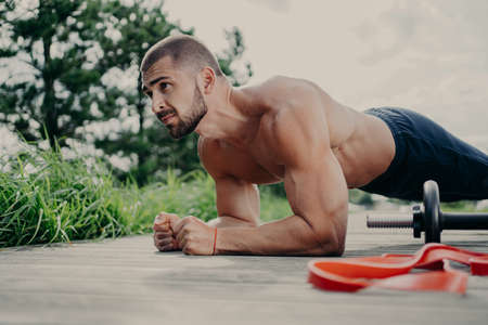 Photo of motivated bodybuilder does plank exercise, stands on elbows, uses sport equipment for workout, concentrated into distance, has muscular body, enjoys training outdoor. Morning fitness conceptの写真素材