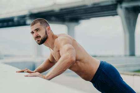 Outdoor shot of bearded muscular man does push up exercises, warms up before training, poses against bridge background, has workout early in morning, concentrated somewhere. Active lifestyleの写真素材