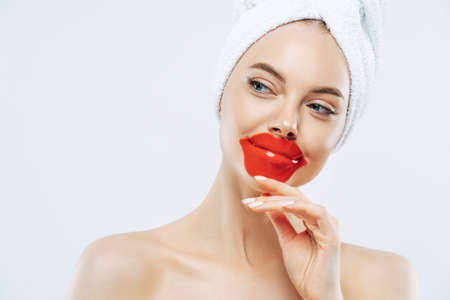Thoughtful beautiful woman wears lip patch, touches jawline gently, has concentrated look aside, enjoys skin care pampering procedure, stands bare shoulders, white background. Cosmetology conceptの写真素材
