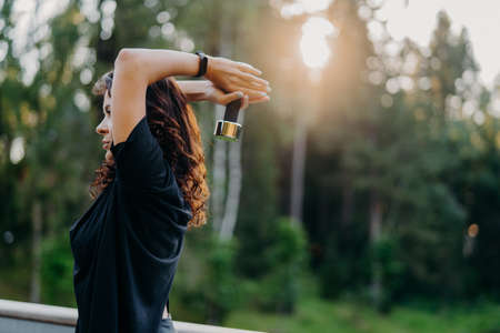 Profile shot of active motivated young woman dressed in black t shirt, raises dumbbell over head, poses against trees with sunset, has workout outdoor, wants to have muscular arms. Sport conceptの写真素材
