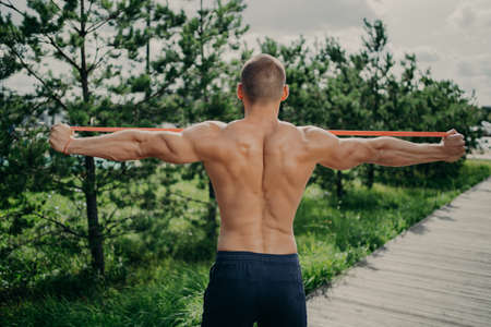 Back view of unrecognizable sportsman stretches elastic resistance band, demonstrares strong muscular body, uses sport equipment, stands outside, enjoys sport exercises. Bodybuilding conceptの写真素材
