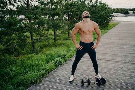 Bearded muscular man keeps hands on waist, raises head and breathes fresh air, takes break after doing workout, trains with barbells. Professional weightlifter works out in nature. Exercising conceptの写真素材