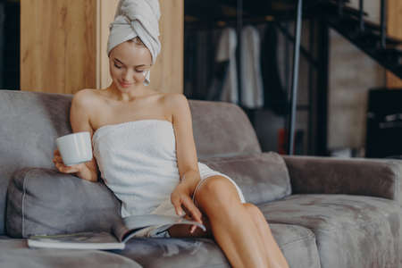 Beautiful young woman wrapped in bath towel, reads beauty magazine, drinks hot beverage, poses on comfortable couch in living room, applies face cream, has healthy smooth skin after taking showerの写真素材