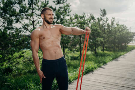 Strength and motivation concept. Muscular man with naked torso, has exercises with resistance band,...