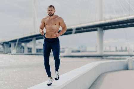 Active bearded male jogger runs along river side has workout outdoor dressed in active wear prepares for sport competitions has muscular strong body breathes fresh air. Sporty lifestyle conceptの写真素材