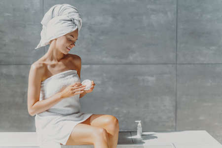 Horizontal shot of relaxed beautiful womam applies body lotion and nourishing cream after shower, poses in bathroom against gray wall, sits wrapped in towel, undergoes beauty treatments. Spa conceptの写真素材