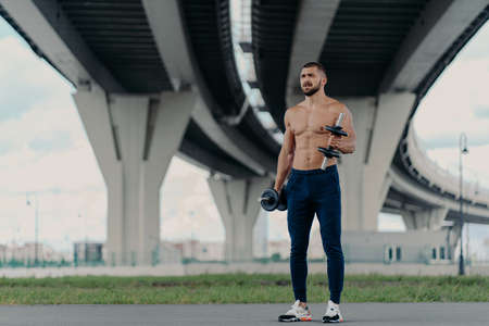Muscular athletic bearded bodybuilder raises dumbbells works on biceps has hard workout near bridge dressed in sport trousers and sneakers, being motivated, enjoys sport. Healthy lifestyle conceptの写真素材