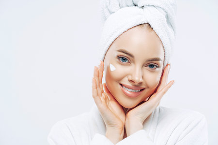Close up portrait of cheerful female with natural beauty, touches cheeks gently, applies face cream for soft skin, wears bath towel on head, dressed in robe, stands indoor. Beauty treatment, lifestyleの写真素材