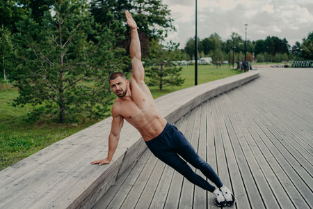 Athletic man stands in side plank pose, raises one arm, poses with shirtless torso, wears trousers...