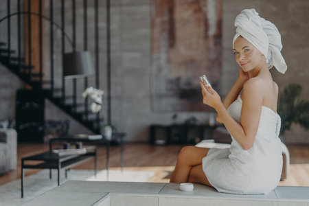 Indoor shot of good looking woman holds bottle of lotion has well cared smooth skin wears towel on head and around naked body after taking shower, poses against home interior. Beauty concept.の写真素材