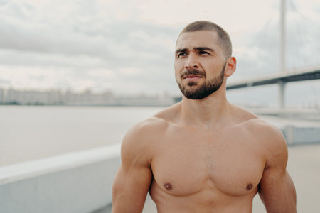 Horizontal shot of muscular man with naked torso thick beard looks somewhere into distance, relaxes outdoors, stands against river background, takes break after jogging, has athletic body shapeの写真素材