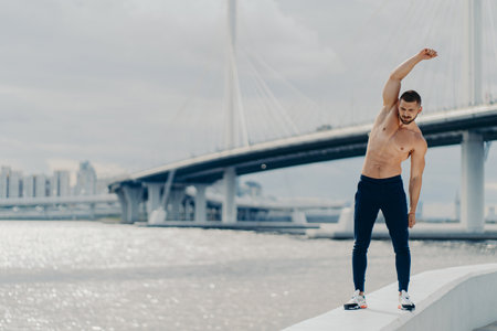 Full length shot of muscles active man does sport exercises outdoor poses near river bridge wears trousers and sneakers has beautiful male torso achieves best shape. Healthy lifestyle conceptの写真素材