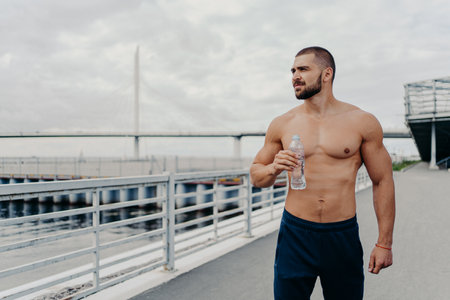 Handsome bearded sportsman drinks water from bottle promots healthy lifestyle and concentrated away into distance, relaxes after morning run, poses outside near river, being thirsty and tired.の写真素材