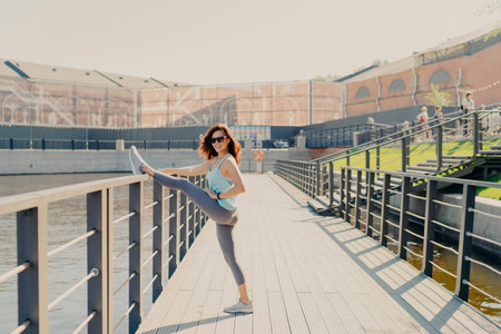Glad brunette woman leans at fence stretches legs has perfect figure dressed in activewear poses...