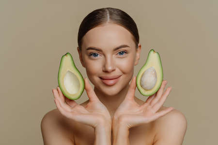 Personal care and spa concept. Brunette satisfied European woman holds halves of avocado near face...