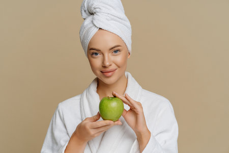 Lovely young woman has refreshed skin after taking bath dressed in dressing gown wrapped towel on head holds green apple containing much vitamins isolated over brown background. Hygiene conceptの写真素材