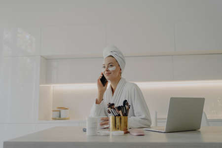 Young attractive business woman in bathrobe and towel turban on head with cosmetic patches under...