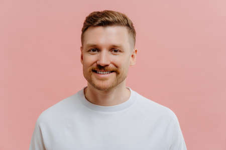 Portrait of handsome pleasant unshaven redhead man expressing positiveness while looking at camera with smile, happy guy in white tshirt feeling joyful while posing against pink studio background.の写真素材