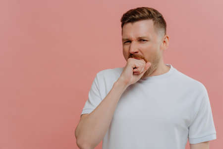 Young bearded man in white shirt yawning and being bored or tired of talk with somebody, sleepy guy covering opened mouth with fist while standing isolated over pastel pink studio backgroundの写真素材