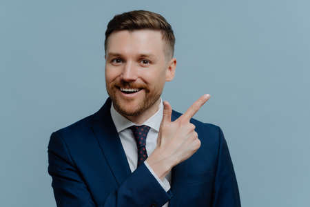 Happy positive businessman in suit pointing with his finger away at copy space and smiling at camera, generating business idea or solution while standing against light steel blue backgroundの写真素材