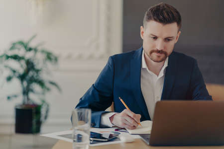 Successful attractive businessman in formal dark blue suit writing down important data about his business project into notepad while sitting at his work desk in front of laptop in stylish officeの写真素材