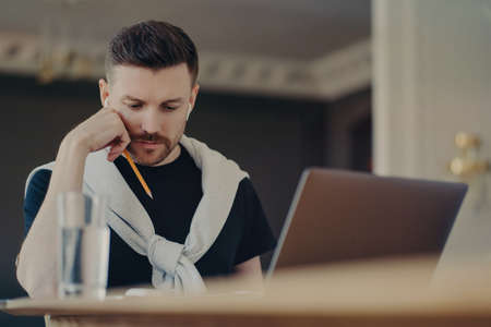 Concentrated male freelancer in wireless earphones and casual clothes being focusd on work, holding...