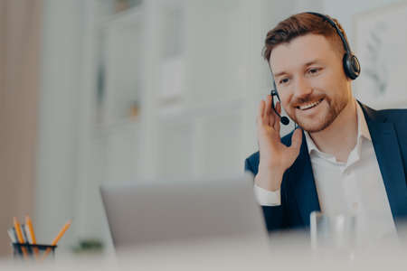 Smiling bearded businessman touching his hand on headset while having business meeting online, male...