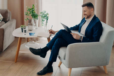 Side view of smiling bearded businessman in suit watching online business video while sitting in armchair and working remotely, using laptop and studying online. Business people lifestyle conceptの写真素材