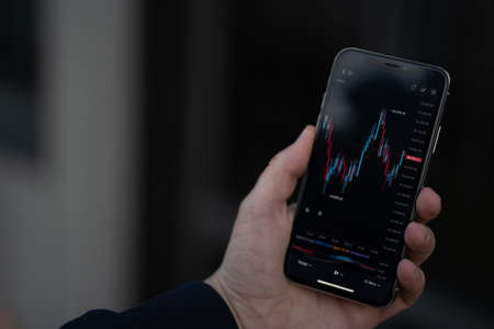 Mobile app for trading online. Male hand holding smartphone with financial forex graph chart on...