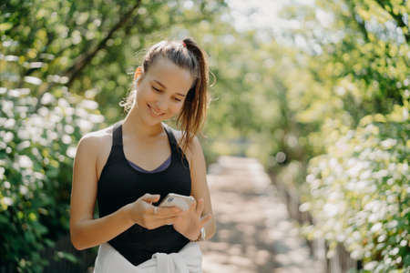 Outdoor shot of happy athletic young woman uses smartphone sport tracker application uses modern device for health and fitness being smart about training poses against blurred nature backgroundの写真素材