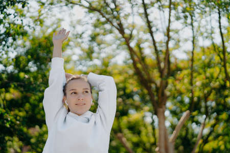 Relaxed beautiful young woman does stretching exercises outdoors keeps arms raised up focused into distance wears white hoodie listens music in earphones has training in park during sunny dayの写真素材