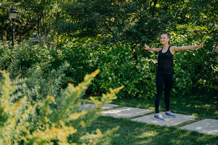 Pleased active woman streatches arms sideways warms up before morning workout wears black...