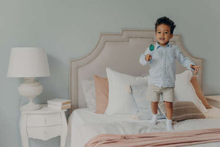 Happy playful afro american child boy with lollipop having lots of fun in bedroom, jumping on bed...