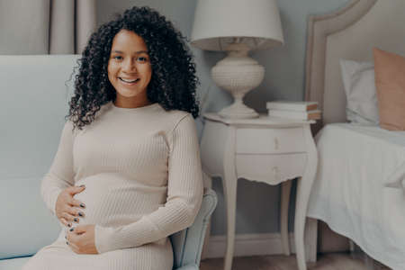 Portrait of young positive pregnant woman sitting in bedroom and looking at camera with bright smile, holding her belly and enjoying pregnancy time while relaxing on armchair at homeの写真素材