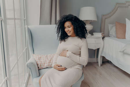 Charming young mixed race pregnant woman, future mother looking out of big window and dreaming about maternity while sitting sideways on armchair in bedroom, holding belly with both hands and smilingの写真素材