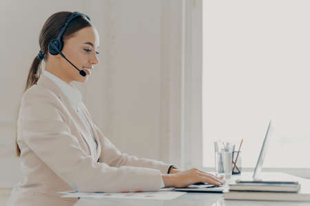 Young friendly female operator in beige formal suit wearing headset and talking with...